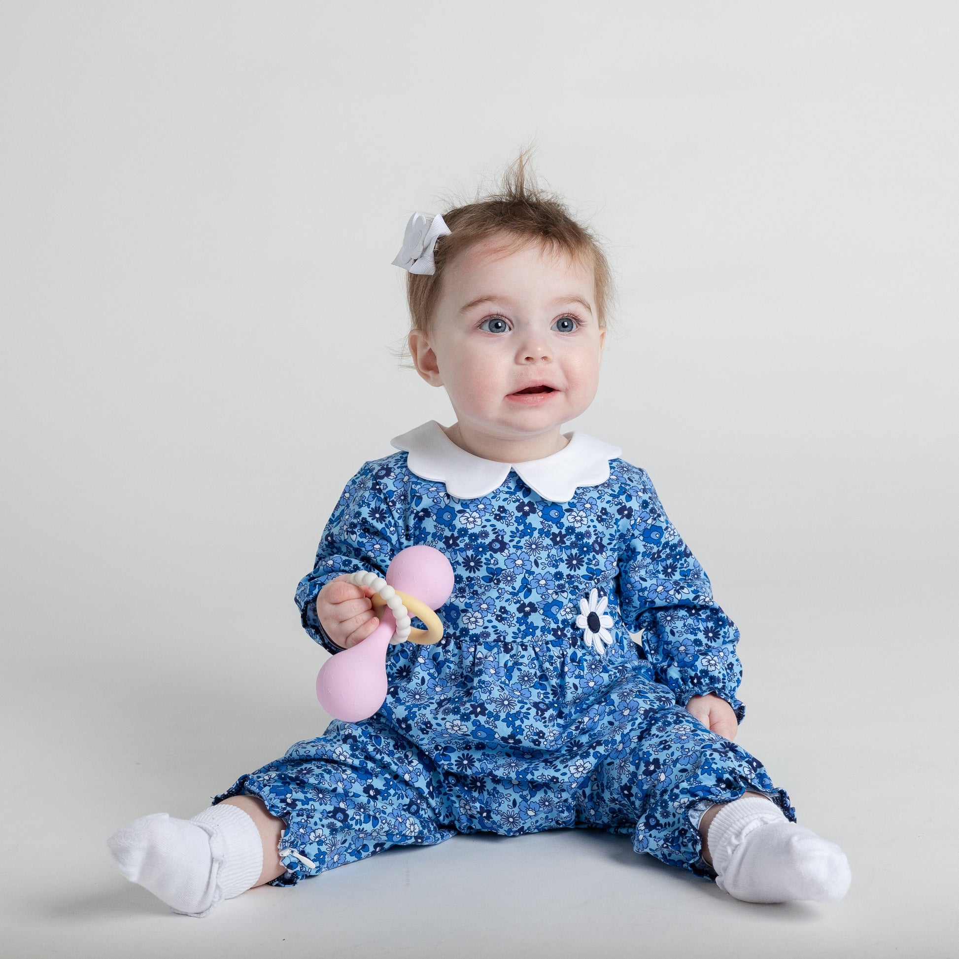 Baby in a blue floral romper holding a pink rattle on a light gray background